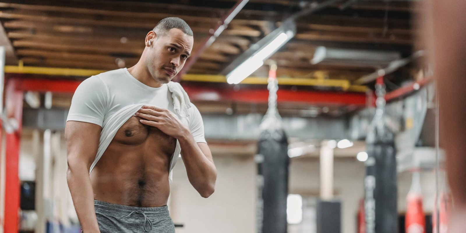 Homem em uma academia, levantando a camiseta para mostrar o torso musculoso enquanto se olha no espelho.